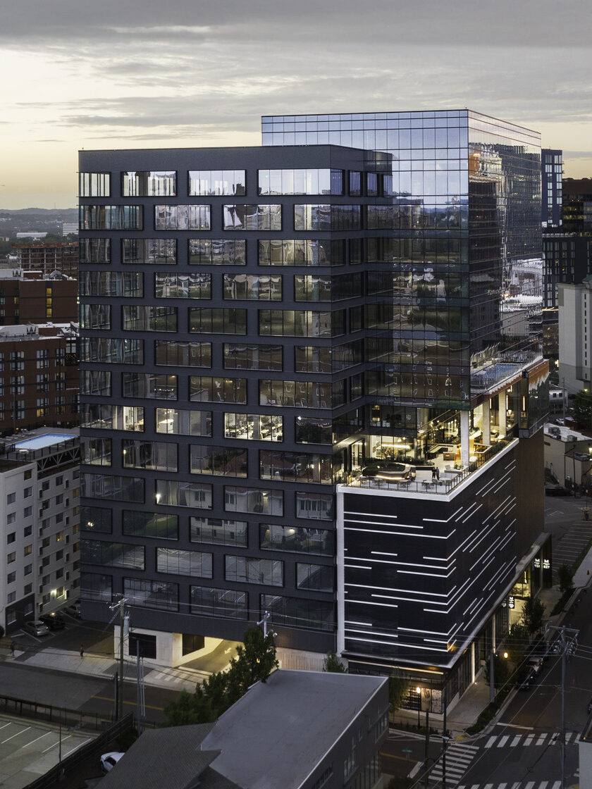 An external view of a downtown building at sunset. The building is covered in glass windows and has a terrace midway up on the right with outdoor seating.