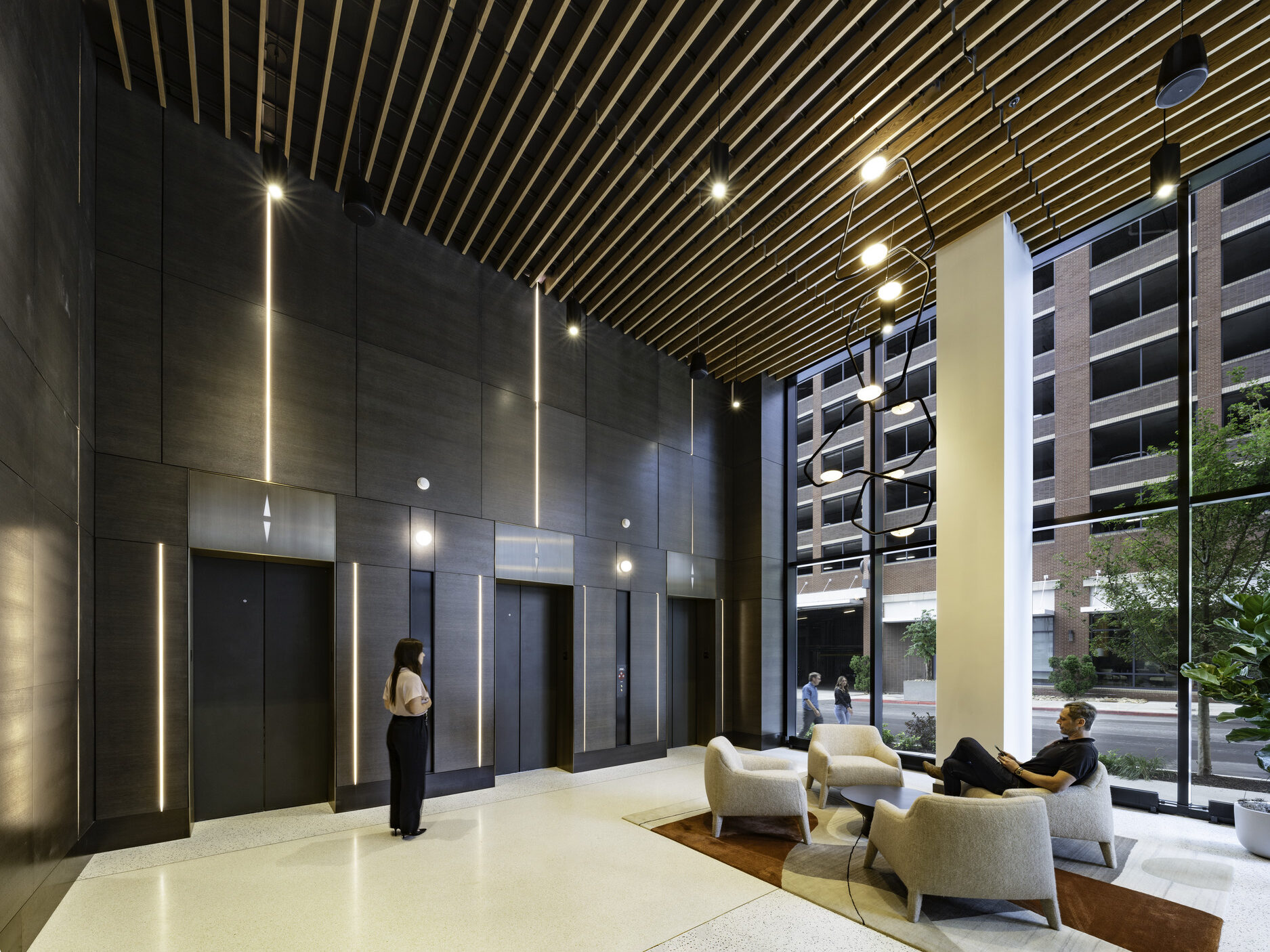 An elevator lobby with a wall of windows to the right and the elevators straight ahead. A woman waits for an elevator as a man sits in a chair in the lobby.