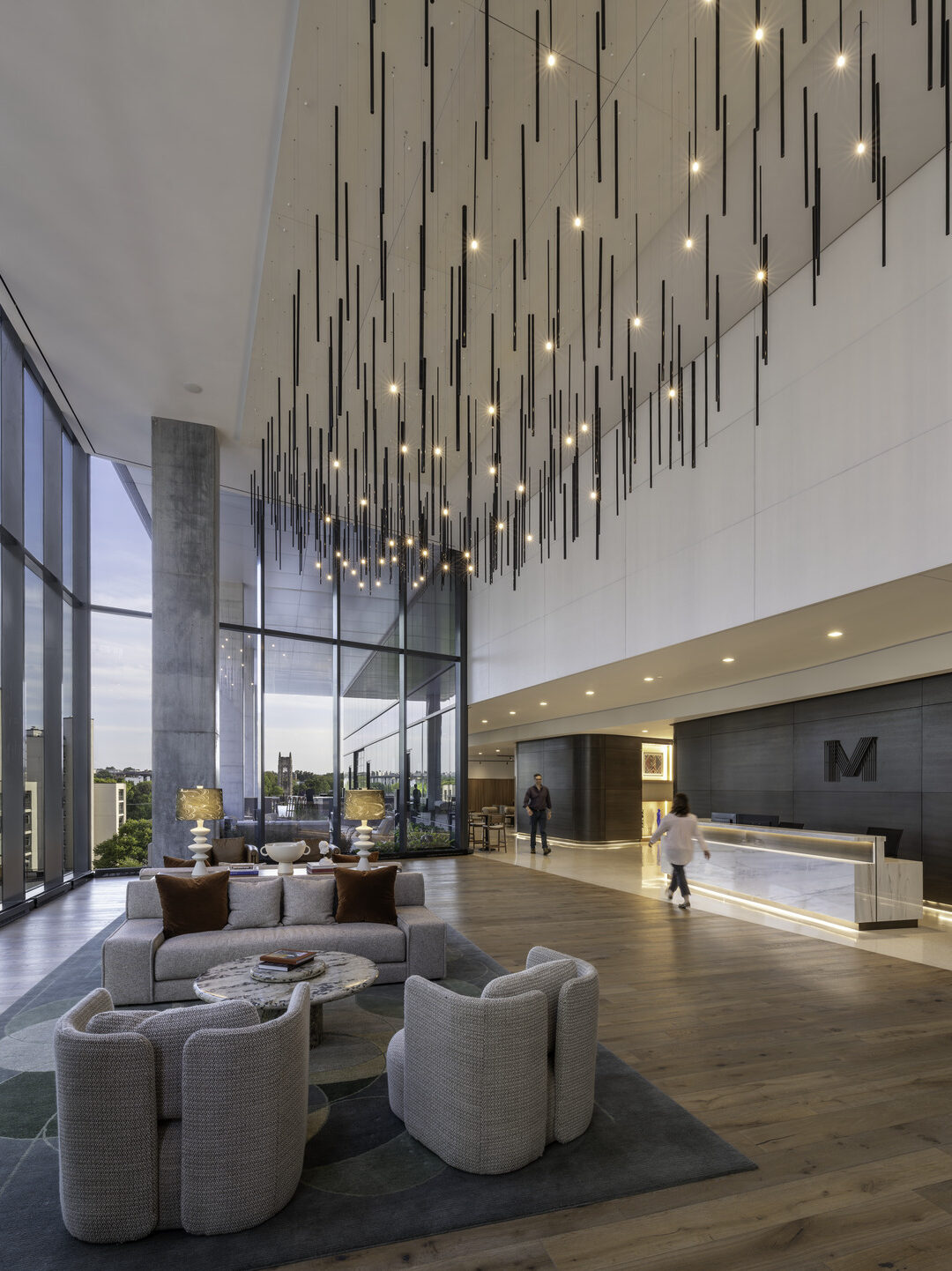 An office lobby with a wall of windows on the left with a check in desk on the right and soft seating in the middle under a modern hanging lightbulb chandelier.