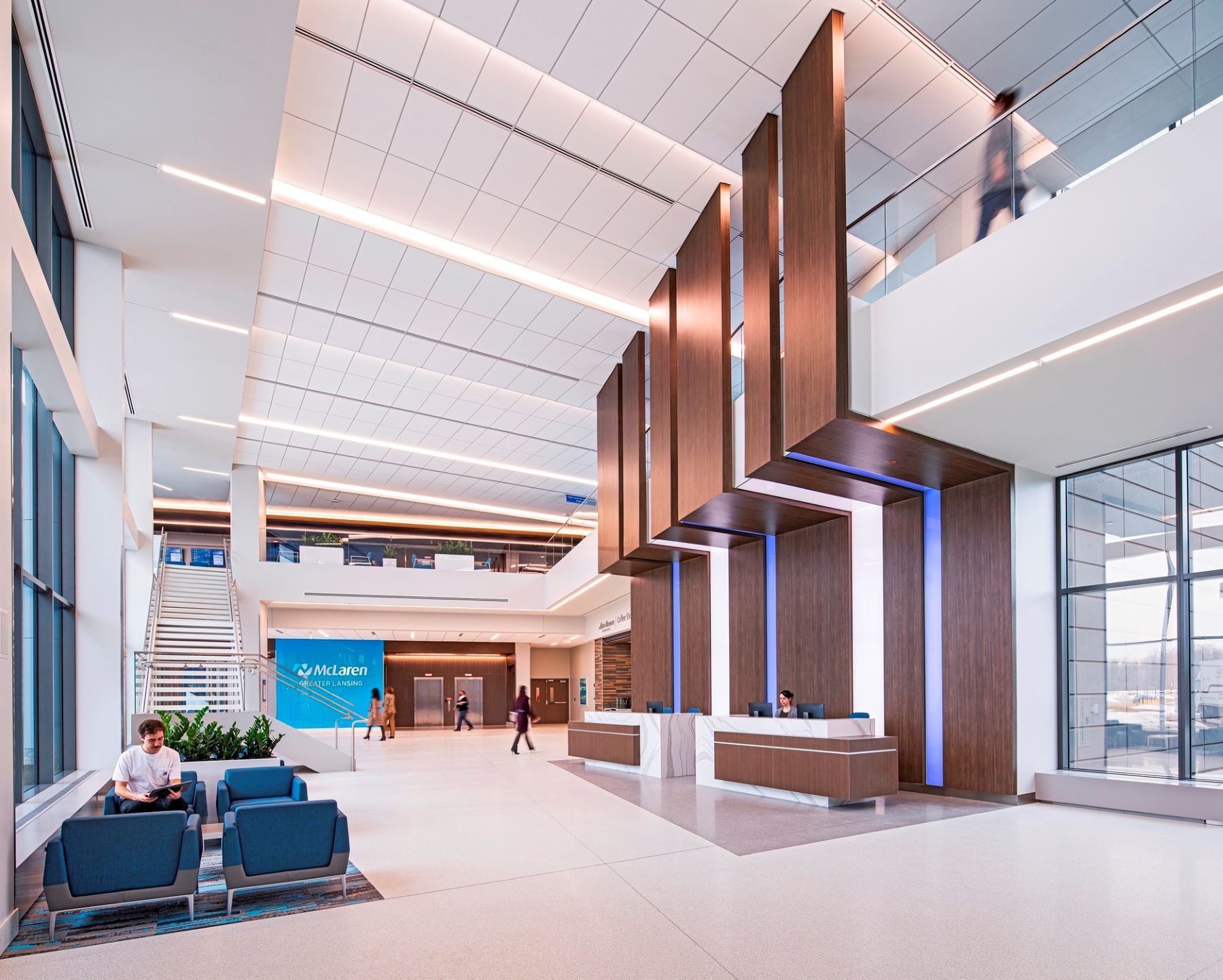 A person sitting in a chair, a person behind a reception desk, and four people walking in a two-story lobby of a hospital