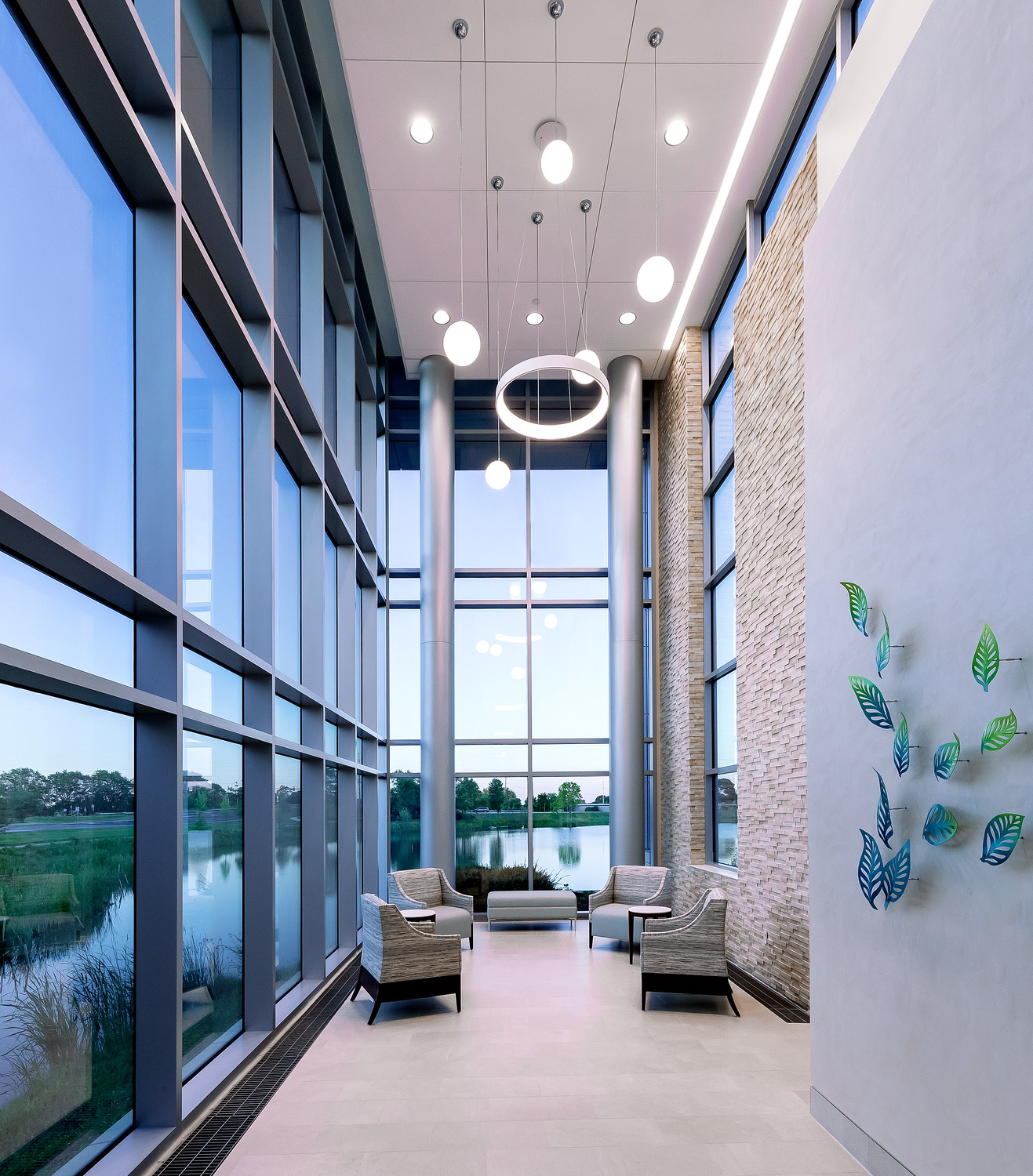 A corridor with two-story ceilings and large windows overlooking a pond