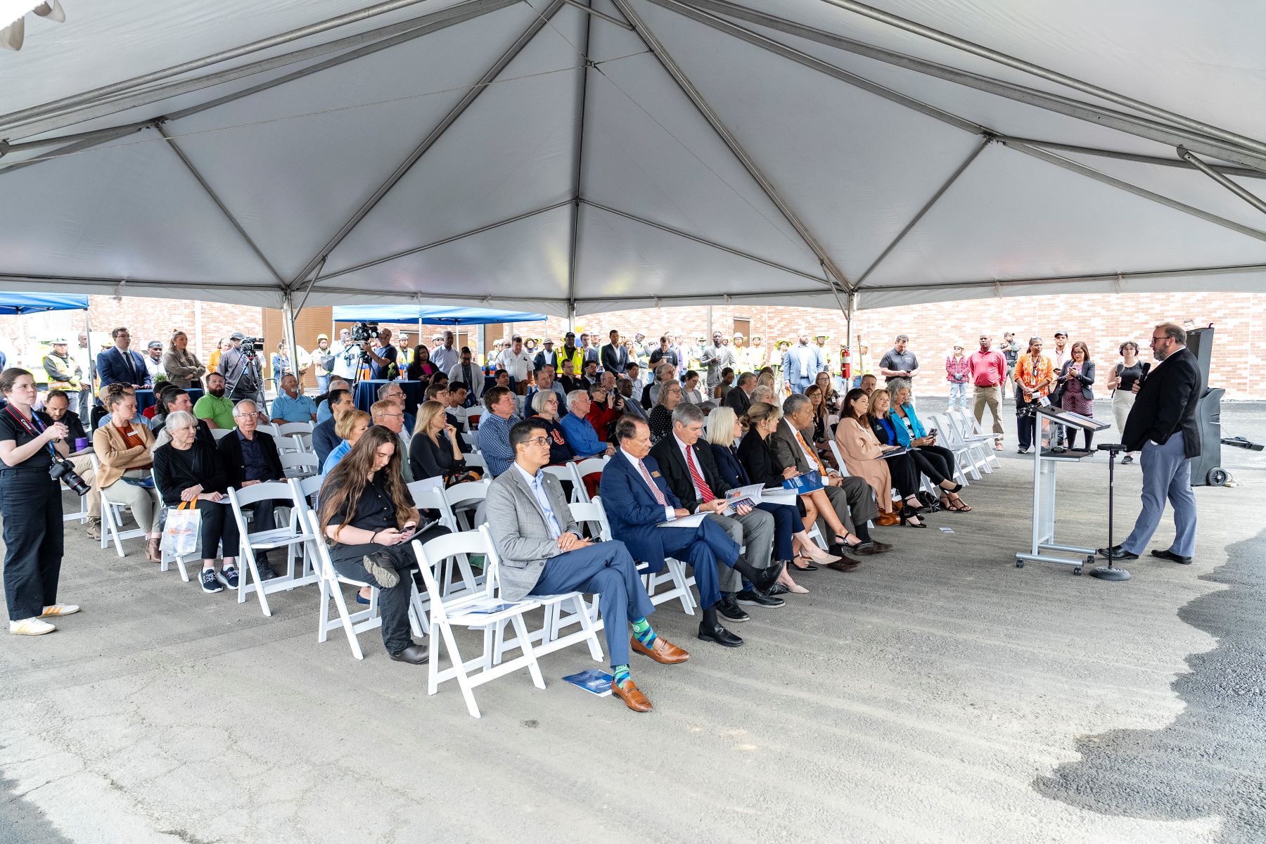 people sitting under a large tent listening to a speaker at a ribbon cutting ceremony